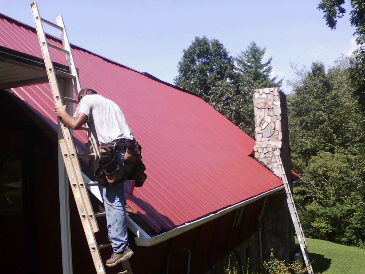 Roofer on a ladder beside a red metal roof and stone chimney — Soffit & Fascia Repair by Roofing Brothers in Prompton
